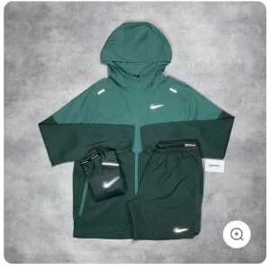 Nike vindjacka med huva och dragkedja - Nike vindjacka med huva och hel dragkedja framtill. Jackan finns i flera färgkombos: mörkgrön, rosa, marinblå och ljusgrön, alla med kontrastfärgade ärmar och reflekterande detaljer. Perfekt för träning eller när du vill ha en sportig vibe. Lätt material och relaxed fit.vi har vilken storlek som helst