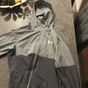 Grå vindjacka från Under Armour - Snygg tvåfärgad grå vindjacka från Under Armour med huva och dragkedja hela vägen. Jackan har en sportig look, logga på bröstet och praktiska fickor. Perfekt för blåsiga dagar och outdoor-aktiviteter.