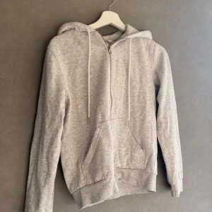 Ljusgrå hoodie med dragkedja från H&M - En ljusgrå hoodie med dragkedja framtill och huva med snören. Tröjan har känguruficka och ribbade muddar vid ärmen och kanten där nere.