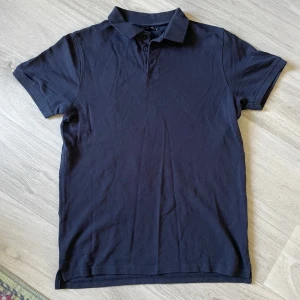 Mörkblå pikétröja från 157 - Snygg mörkblå pikétröja från 157 med klassisk krage och knappar framtill. Tröjan är i mjuk bomull och har korta ärmar, perfekt för en clean och stilren look. Passar dig som gillar enkel och tidlös stil.
