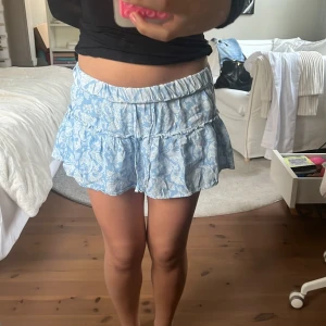 Blå volangkjol från Zara - Supersöt blå kortkjol från Zara med vitt mönster. Går att vika ned för lägre midja!! Har insydda shorts. Använt fåtal gången och är i jättebra skick!