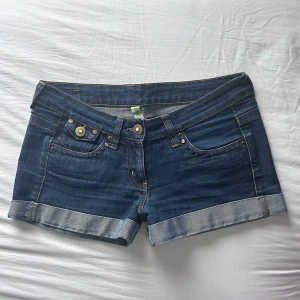 Lågmidjade jeansshorts - Snygga blå jeansshorts med klassisk femficksdesign. Shortsen har knappar och nitar samt bakfickor med lock och knappdetalj. Perfekta för varma dagar och enkel att matcha med allt. Storlek 38, dvs M