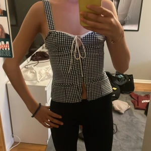 Rutig korsettopp från Zara - Supersöt svartvit rutig korsettopp från Zara med breda axelband och knytning framtill. Toppen har en figurnära passform och är perfekt för dig som gillar en trendig och feminin look. Tillverkad i bomullsmix med snygga detaljer vid bysten.