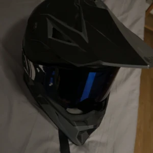 Svart motocrosshjälm med visir - Säljer en svart motocrosshjälm med coolt blåtonat visir och vit-svart grafiskt mönster på sidorna. Hjälmen har en skärm framtill och sportig design, perfekt för dig som gillar fart och äventyr. Justerbart hakband med röd detalj.
