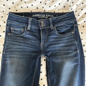 Lågmidjade Mörkblå jeans från American Eagle - Snygga mörkblå jeans från American Eagle med klassisk femficksdesign och dubbla knappar i midjan. Jeansen har raka ben och lätt slitna detaljer för en cool look. Materialet är bomull med lite stretch för extra komfort. Lågmidjade. Avklippa i benen passar mig som är ca 160