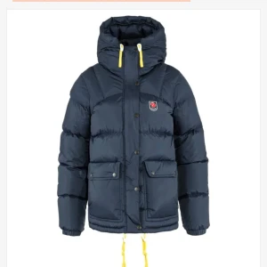 Mörkblå dunjacka från Fjällräven - Säljer en mörkblå dunjacka från Fjällräven med huva och gula snören. Jackan har två stora fickor framtill, dragkedja och knappar. Perfekt för kalla dagar och har en klassisk Fjällräven-logga på bröstet. Snygg och funktionell stil.