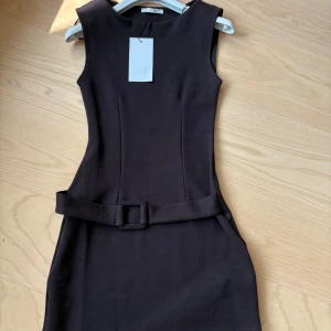 Brun klänning från Zara nyskick - Snygg svart ärmlös kort klänning från Zara med markerad midja och matchande bälte. Klänningen har en stilren, figurnära passform och rund halsringning. Helt ny aldrig använd med prislapp!! 