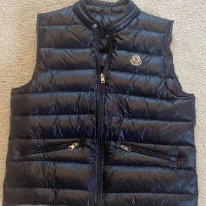 Mörkblå dunväst från Moncler - Snygg mörkblå dunväst från Moncler med glansig finish och klassisk logga på bröstet. Västen har två dragkedjefickor framtill och hög krage. Perfekt att slänga över en hoodie eller t shirt inför sommaren. NFC tag på både loggan och på etikettten.