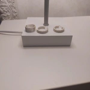 Silvrigt set med ring och örhängen - Säljer ett snyggt set med en bred silverfärgad ring och ett par matchande creolörhängen. Både ringen och örhängena har ett vågigt, texturerat mönster som ger en cool och modern vibe. Perfekt för dig som gillar statement-smycken i stilren design.Silver 925.