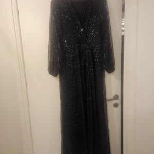 Mörkblå glittrig långklänning Angeleye - Säljer en mörkblå långklänning från Angeleye med glittriga paljetter i ränder över hela klänningen. Djup v-ringning, långa ärmar och lätt transparent tyg med fodrad insida. Perfekt för dig som vill sticka ut med en elegant look.