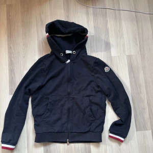 Svart Moncler hoodie med huva - Mörkblå hoodie från Moncler med dragkedja, huva och fickor framtill. Ärmsluten har vita och röda ränder för en sportig look. Moncler-logga på ena ärmen. Perfekt för dig som gillar stilrena och exklusiva plagg. Mycket bra skick. Hör av er vid frågor 