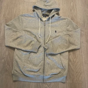 Grå hoodie från Polo Ralph Lauren - Snygg ljusgrå hoodie från Polo Ralph Lauren med dragkedja och vit snörning i huvan. Klassisk broderad logga på bröstet och ribbade muddar. Tillverkad i mjuk bomullsmix, perfekt för chill dagar eller när du vill ha en clean look.