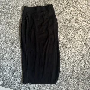 Svart ribbad långkjol med slits - Svart långkjol med ribbad struktur och en snygg slits på sidan. Kjolen har en rak passform och är perfekt för dig som gillar stilrena och enkla plagg. Materialet känns mjukt och följsamt mot kroppen.