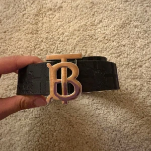 Svart Burberry bälte med TB-spänne - Snyggt svart bälte från Burberry med klassiskt rutmönster och präglad logga. Bältet har ett stort metallspänne med TB-monogram i silverfärg. Perfekt accessoar för att lyfta din outfit med en lyxig touch.