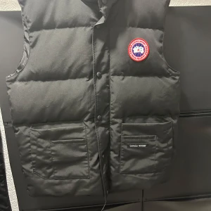 Svart dunväst från Canada Goose Storlek L - Säljer en svart dunväst från Canada Goose med klassisk logga på bröstet. Västen har två stora fickor framtill, tryckknappar och en stilren, quiltad design. Perfekt för lager-på-lager och håller dig varm under kyliga dagar.