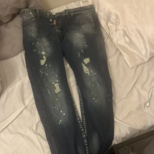 Dsquared2 ICON jeans med färgstänk - Mörkblå Dsquared2 jeans med slitningar, färgstänk och stor vit 'ICON'-patch bak. Klassisk femficksmodell i bomull med lite stretch. Snygg streetstil och coola detaljer framtill. Perfekta för dig som gillar statement-plagg.