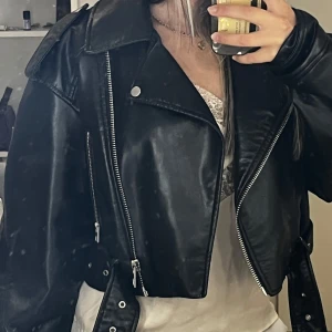 Svart skinnjacka från Zara, croppad - Säljer en svart croppad skinnjacka från Zara med coola silvriga dragkedjor, bred krage och bälte i midjan. Jackan har en oversized passform och långa ärmar, perfekt för att lyfta vilken outfit som helst.