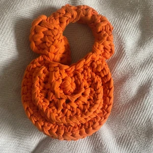 Handgjord orange stickad handväska - märke Li Per Leí - Unik rund handväska i starkt orange färg, stickad med grovt garn för en chunky look. Väskan har ett matchande handtag och kommer med en liten orange påse i satin. Perfekt för dig som vill sticka ut med accessoarer.