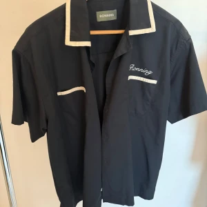 Svart skjorta från Ronning med kontrastdetaljer - Svart skjorta från Ronning med kort ärm och vita kontrastdetaljer på krage och bröstfickor. Broderad logga på bröstet. Skjortan är tillverkad i en mix av polyester och bomull, vilket gör den både mjuk och lättskött. Perfekt för en clean och stilren look.