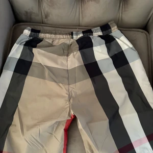Burberry rutig shorts med snörning - Snygga Burberry shorts i klassiskt rutigt mönster med beige, svart, vit och röda detaljer. De har elastisk midja och vit snörning för skön passform. Perfekta för sommaren och ger en trendig vibe till din outfit.