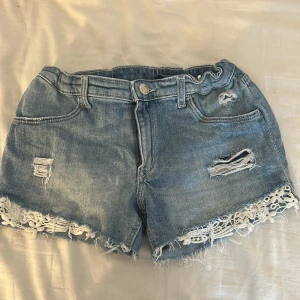 Blå jeansshorts med spetsdetalj - Snygga blå jeansshorts med slitna detaljer och råa kanter. Nederkanten är dekorerad med vit spets som ger en extra touch. Verkligen jätte fina short men är tyvärr för små! 