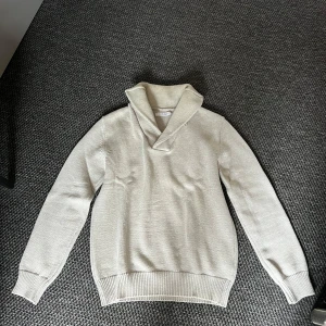 Beige stickad tröja Our Legacy - Beige stickad tröja från Our Legacy med bred krage och bruna mockadetaljer på armbågarna. Tröjan har lång ärm, ribbade muddar och en clean, stilren look som passar perfekt till lager-på-lager. Snygg och enkel att matcha med jeans eller chinos.