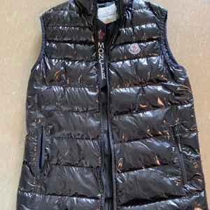 Svart dunväst från Moncler, S - Snygg svart dunväst från Moncler med glansig finish och klassisk logga på bröstet. Västen har dragkedja framtill, två fickor med dragkedja och quiltad design. 
