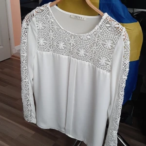 Vit blus med spets från IVIVI - Superfin vit blus från IVIVI med långa ärmar och detaljerad spets upptill och längs ärmarna. Blusen har rund halsringning och ett luftigt, flowy tyg som känns lätt på kroppen. Perfekt för dig som gillar romantiska och stilrena vibes.