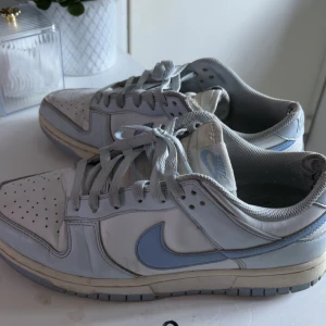 Nike Dunk sneakers i vitt och ljusblått - Nike Dunk sneakers i vitt och ljusblått skinn med klassisk låg siluett. Skorna har perforerad tå, ljusblå swoosh och matchande snörning. Perfekta för dig som gillar retrovibbar och streetstyle. Snygg kontrast mellan de ljusa färgerna och den robusta sulan. jätte använd 