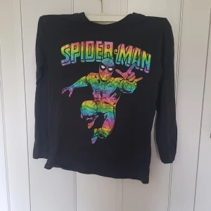 Svart Spiderman-tröja från H&M Marvel - Cool svart långärmad tröja från H&M Marvel med färgglatt Spiderman-tryck på bröstet. Tröjan har en klassisk passform och är gjord i mjuk bomull, perfekt för dig som gillar superhjältar och vill sticka ut med en färgsprakande look.