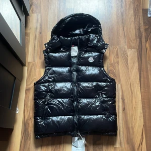 Svart dunväst från Moncler med huva - Snygg svart dunväst från Moncler med glansig finish, avtagbar huva och två dragkedjefickor. Västen har klassisk Moncler-logga på bröstet och grov dragkedja framtill. Perfekt för lager-på-lager och streetstyle.