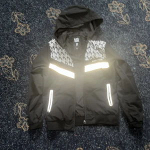Svart vindjacka från Black Squad - Svart vindjacka från Black Squad i storlek XS. Jackan har huva, reflekterande detaljer på bröst och fickor samt vitmönstrade partier upptill. Stängs med dragkedja och har ribbade muddar. Perfekt för dig som vill sticka ut med snygga detaljer.