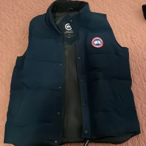 Mörkblå dunväst från Canada Goose - Snygg mörkblå dunväst från Canada Goose ÄKTA! Med klassisk logga på bröstet. Västen har hög krage, tryckknappar och dragkedja framtill. Perfekt för lager-på-lager och håller dig varm under kyliga dagar. Fodrad insida och stilren design.