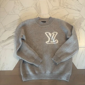 Grå Louis Vuitton stickad tröja - Snygg grå stickad tröja från Louis Vuitton med stort vitt LV-logo framtill. Klassisk rund halsringning och ribbade muddar vid ärmslut och nederkant. Perfekt för dig som vill ha en stilren och exklusiv look. Priset är ej hugget i sten!