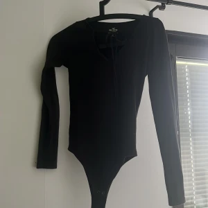 Svart långärmad body från Hollister - Svart långärmad body från Hollister Must-Have Collection. Snygg v-ringning med knyt framtill och smidig knäppning nedtill. Perfekt att styla med jeans eller kjol för en clean och enkel look.