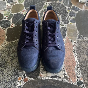 Mörkblå sneakers från Christian Louboutin (42)   - Snygga mörkblå sneakers från Christian Louboutin i mocka med klassisk röd sula och rund tå. Skorna har snörning och diskreta detaljer, perfekt för dig som gillar lyxiga och stilrena sneakers med en twist. Utanskokartong