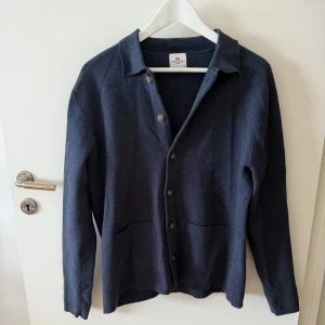 Mörkblå kofta från John Henric - Stilren mörkblå kofta från John Henric med klassisk krage och knäppning framtill. Koftan har två praktiska fickor och är tillverkad i mjukt material som passar perfekt för lager-på-lager. Långärmad och enkel att matcha med olika outfits.