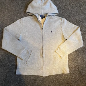 Grå zip hoodie från Polo Ralph Lauren - Snygg ljusgrå hoodie från Polo Ralph Lauren med dragkedja och klassisk broderad logga på bröstet. Tröjan har huva, kjänguruficka.  Perfekt för en chill och stilren look. Storlek 14-16 år. Passar perfekt till folk runt 160-165. Mycket bra skick! Tvättad och klar till utskickning🫧Skriv om ni har funderingar eller prisförslag😃