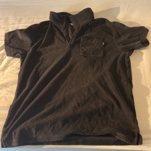 Svart pikétröja i frotté från WRP - Svart pikétröja från WRP i mjuk frotté. Tröjan har korta ärmar, klassisk krage med halv knappslå och en bröstficka. Perfekt för en chill och avslappnad stil under varmare dagar.