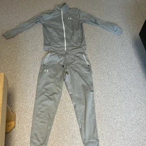Grå mjukisbyxor från Under Armour - Snygga grå mjukisbyxor från Under Armour med dragsko i midjan och vita dragkedjor på fickorna. Byxorna har en slim passform och är tillverkade i ett mjukt syntetmaterial som passar perfekt till träning eller chill. Diskret logga på låret. Koftan S byxorna M