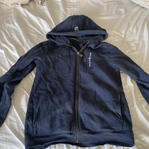 Mörkblå hoodie från Sail Racing - Säljer en mörkblå hoodie från Sail Racing med dragkedja och huva. Den har långa ärmar och en broderad logotyp på bröstet. Perfekt för en avslappnad stil.