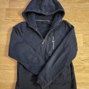 Marinblå Zipp hoodie från Sail Racing - Säljer en marinblå hoodie från Sail Racing med dragkedja och huva. Jackan har en logotyp på bröstet och är tillverkad i ett mjukt material. 