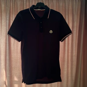 Svart kortärmad pikétröja från Moncler - Snygg svart pikétröja från Moncler med klassisk krage och knappar. Tröjan har en broderad Moncler-logga på bröstet och kontrasterande ränder på krage och ärmslut. Perfekt för en stilren look. Knappt använd