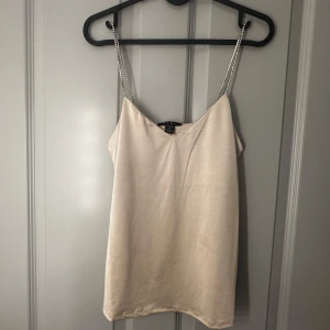 Beige linnetopp med kedjeband - Snygg beige linnetopp med glittriga kedjeband som axelband.Toppen har en enkel och stilren design som passar till många olika outfits.