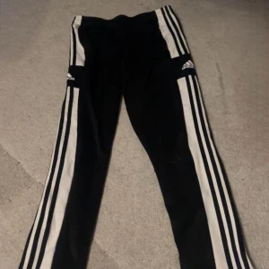 Svarta träningsbyxor från Adidas - Snygga svarta träningsbyxor från Adidas med de klassiska tre vita ränderna längs sidorna. Byxorna har dragkedja vid bensluten och är tillverkade i Primegreen-material. Perfekta för sport och träning. Super sköna och stretch i midjan. Sitter som en smeck, skit bra pass form.
