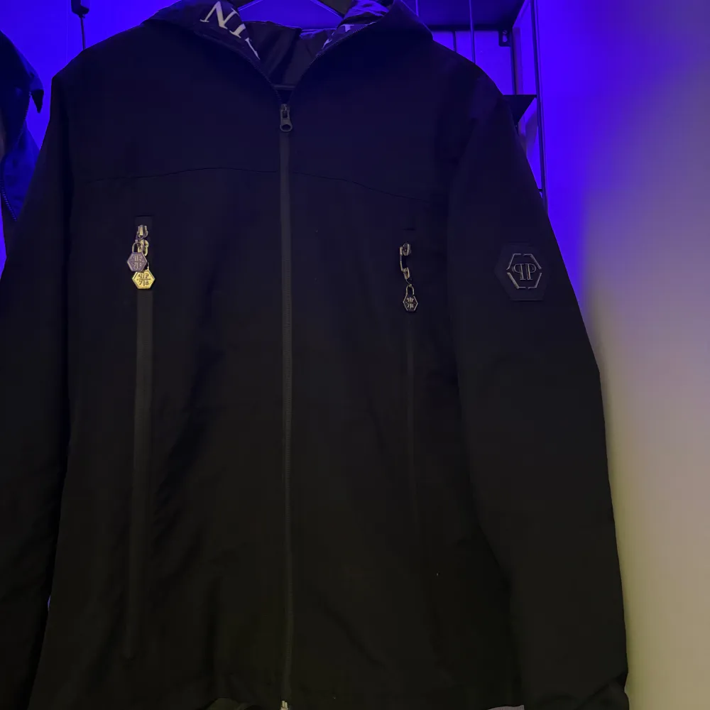 Snygg svart jacka från Philipp Plein med dragkedja och huva. Jackan har märkets logga på ärmen och text på insidan av huvan. Perfekt för en stilren look.. Takit.