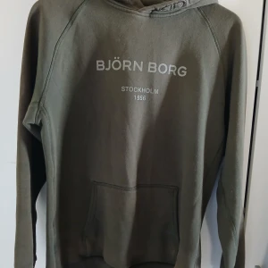 Olivgrön hoodie från Björn Borg - Olivgrön hoodie från Björn Borg med tryck på bröstet med klassisk känguruficka och en bekväm huva. Finns några noppror som syns på bilder. 