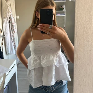 Vit ärmlös blus med spetsdetaljer - Säljer en söt vit ärmlös blus med smala axelband och spetsdetaljer. Blusen har ett volanglager och ett broderat blommönster som ger en romantisk känsla. Perfekt för varma sommardagar de är storlek 170 men skulle säga s-m.