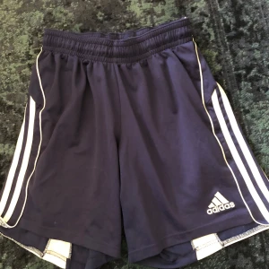 Mörkblå shorts från Adidas - Säljer ett par mörkblå Adidas shorts med vita detaljer och klassiska ränder på sidorna. Perfekta för sport och träning. Elastisk midja för bekväm passform.