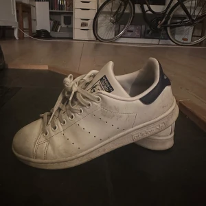 Vita Adidas Stan Smith sneakers - Klassiska vita Adidas Stan Smith sneakers med blå detaljer. Skorna har snörning och är tillverkade i syntetmaterial. Perfekta för en stilren och tidlös look.